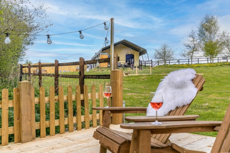 a glass of rose wine on a wooden table next to a wooden chair with a white fur throw. in the background is a cream canvas safari tent and a blue sky. unique places to stay in devon.