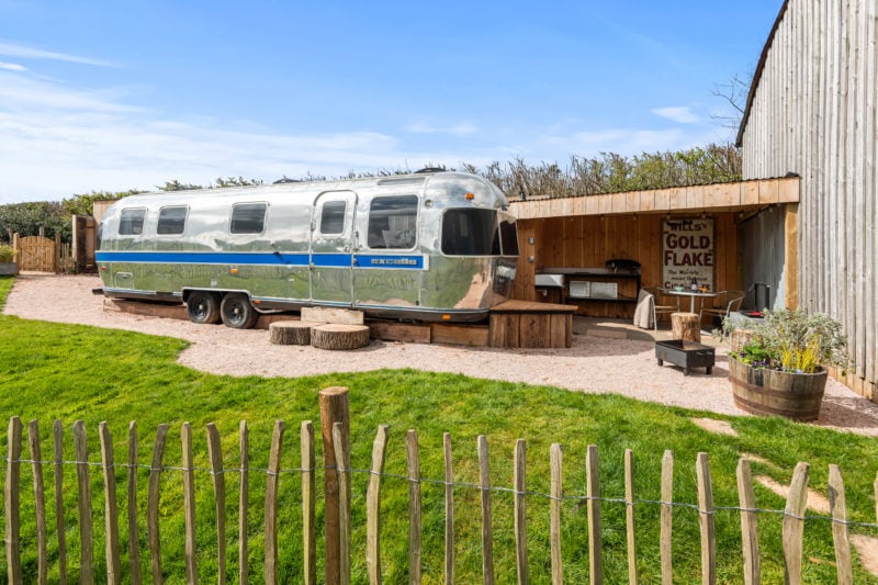 a silver metal airstream caravan in front of a grassy lawn with the edge of a wooden barn to the right. unique places to stay in devon. 