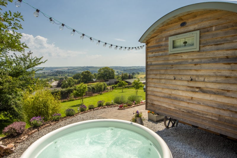 a hot tub next to the edge of a wooden shepherds hut with a view of the green Devon countryside behind.