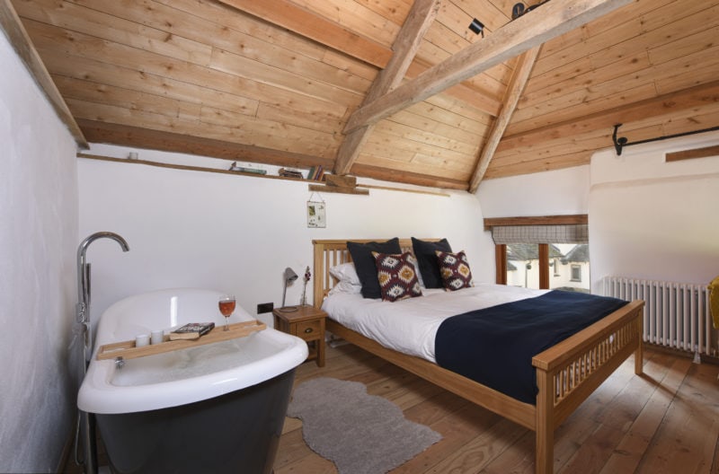 interior of a log cabin with a cieling made of light brown pine and walls painted white. there is a wooden bed with white and blue covers and a free standing bath tub in the room. unique places to stay in devon.