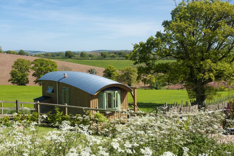 a small brown wooden wagon with green painted windows and a curved grey roof in front of green fields and trees.