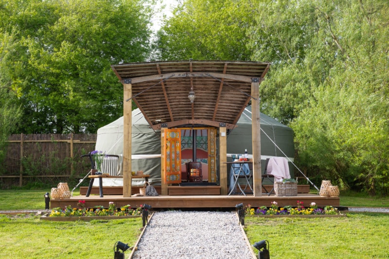 a circular yurt style tent made of white canvas with an open door. in front there is wooden decking and behind are green trees.