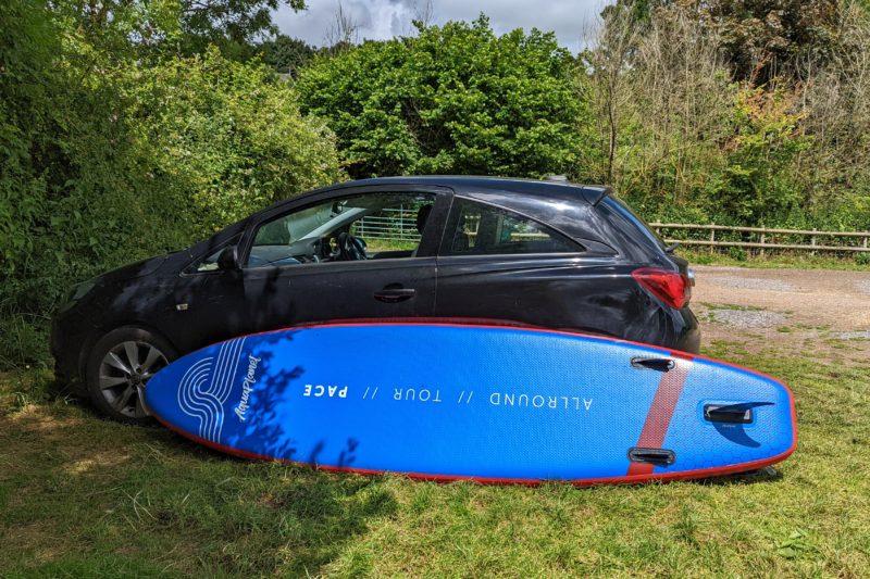 a blue SUP board leaning against a black Vauxhall Corsa parked in a grassy field alongside some bushes