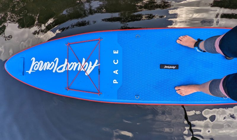 looking down at a bright blue paddleboard on water with a girl's legs in black and grey leggings. aquaplanet pace review