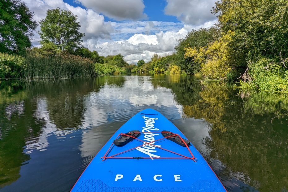 front nose of a bright blue paddleboard on a river with trees and a blue sky behind it, reflected in the river. aquaplanet PACE SUP review.