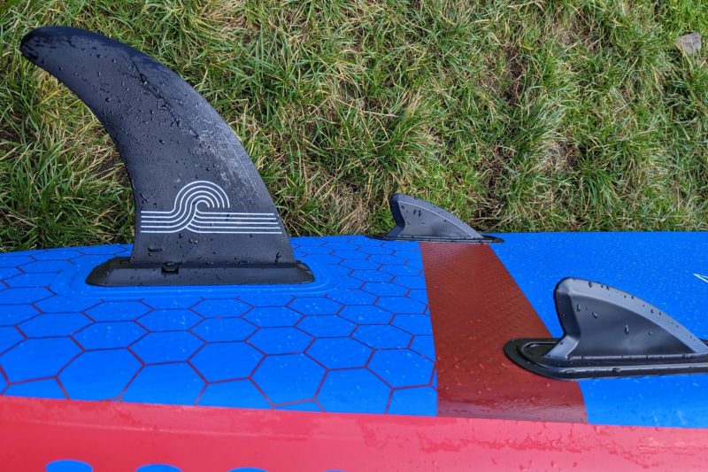 three black fins protruding from the back of a bright blue paddleboard (side view) with grass beneath it. Aquaplanet PACE review.