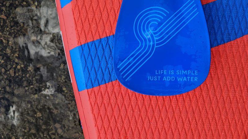 close up of a blue paddle on the edge of a red paddleboard. the paddle has a swirled design on it and the words "life is simple just add water"