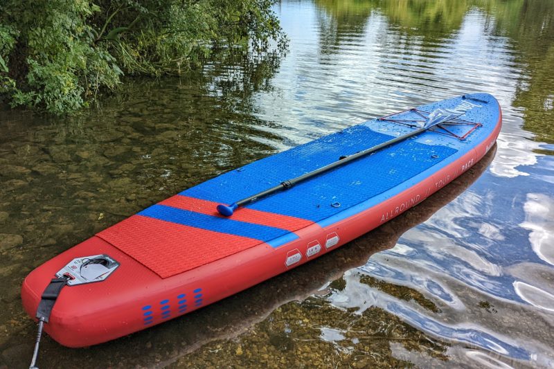 a red and blue paddleboard with a black paddle lying on top floating in a river with brown water and some bushes just visible at the edge of the frame. Aquaplanet PACE review. 