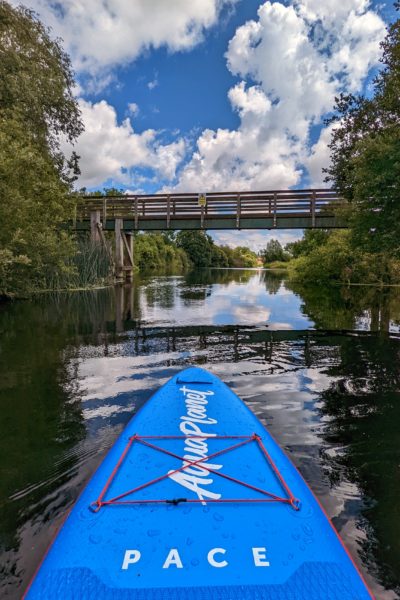 front nose of the Aquaplanet PACE paddleboard, bright blue with the word PACE in white across it, on a river heading towards a wooden bridge with a blue sky behind it. 