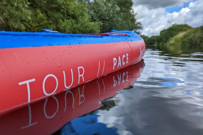 close up view of the side of a paddleboard with a red edge and the words 'tour pace' written in white along the side. the top of the board is blue and the board is on a river with green bushes trees out of focus behind it.