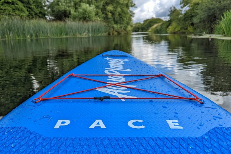 Close up of the nose of a bright blue paddle board with red bungee cords, with the words "aquaplanet pace" printed in white. The board is on a river with out of focus reeds on either bank and trees in the distance. 