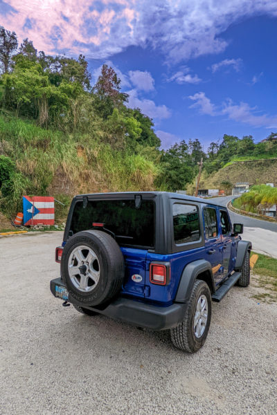 a blue jeep seen from the back on a gravel drive with a puerto rico flag against a small cliff covered in grass and tropical plants in the background and a mountain road seen to the right of that on a very sunny day with a blue sky