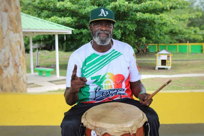 bomba drummer sitting in front of a drum wearing a white t shirt with a green and red logo and a dark green cap with the letter A on it smiling at the camera and giving the peace sign