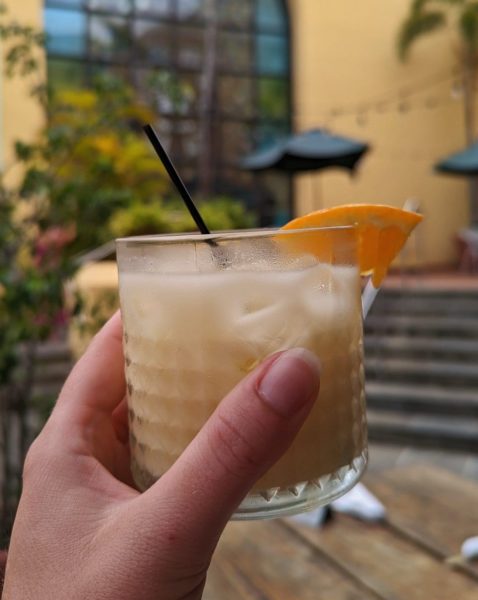 hand holding a yellow cocktail in a short glass tumbler with a slice of orange in front of a yellow wall at a bar in san juan PR