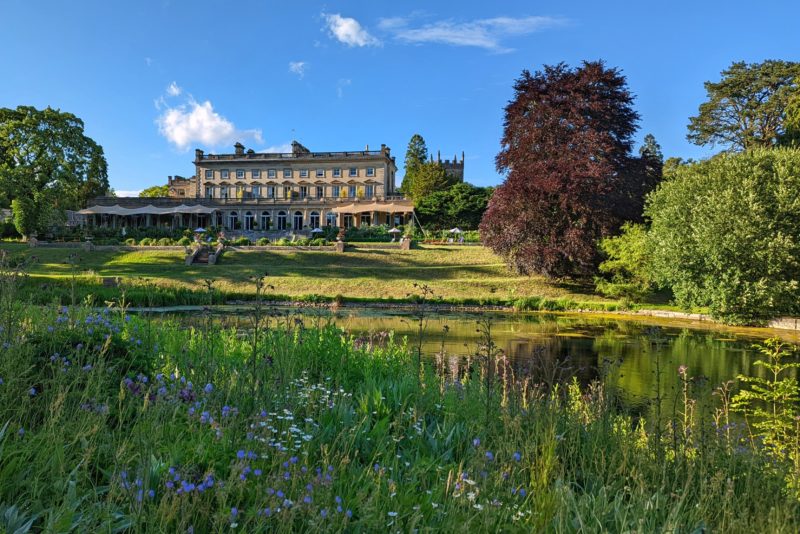 Looking across a lake and a green lawn surrounded by trees towards Cowley Manor Hotel - where to stay near Cheltenham and Gloucester