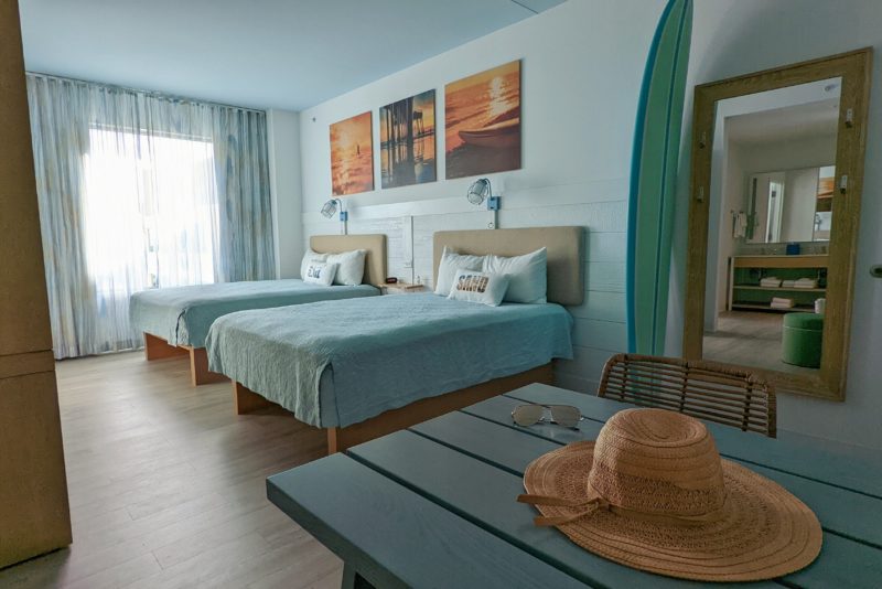 Hotel room painted in pale blue with two double beds with pale cyan bedsheets. there is a wooden blue picnic table in the foreground with a straw hat and sunglasses on it. budget accommodation at universal studios in orlando florida