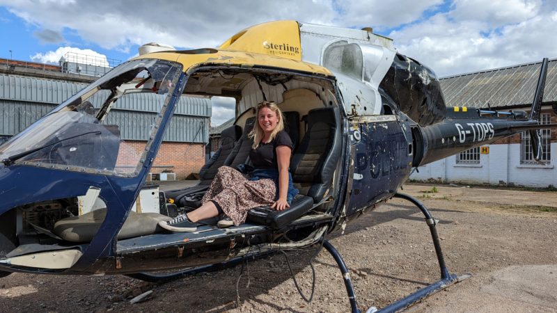 Emily wearing a black t shirt and leopard print skirt sitting in an old broken blue helicopter in Gloucester Prison