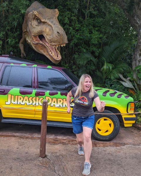 emily wearing denim shorts and a grey t shirt with the jurassic park logo pretending to run towards the camera with a bright green and yellow jurassic park jeep and a life-sized t-rex behind her in universal studios orlando florida 