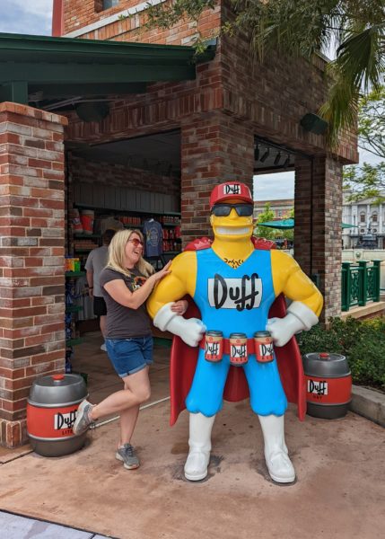 emily wearing denim shorts and a grey t shirt linking arms with a statue of Duff Man from the Simpsons and pretending to laugh while touching his bicep. how to do universal studios orlando on a budget.