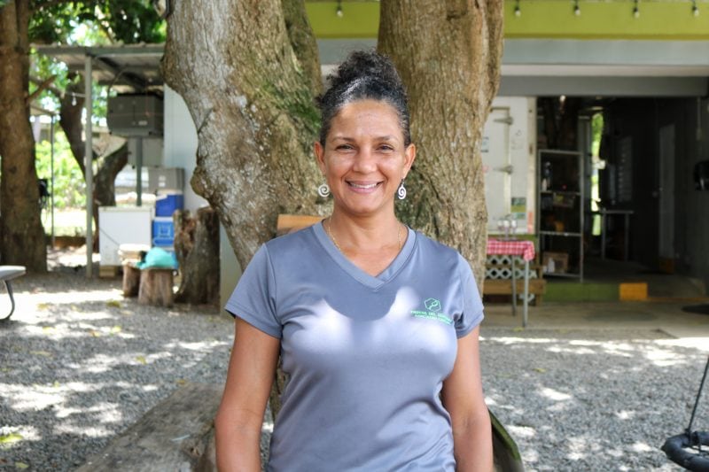 a lady with curly dark hair tied up wearing a grey t shirt standing in front of the camera and smiling in front of her farmhouse in puerto rico
