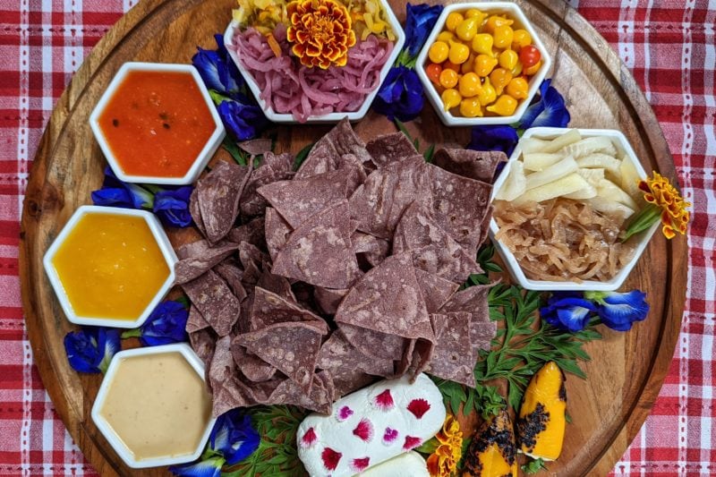 flat lay of several small bowls arranged in a circle around a pile of brown tortilla chips on a wooden tray on top of a red and white table cloth