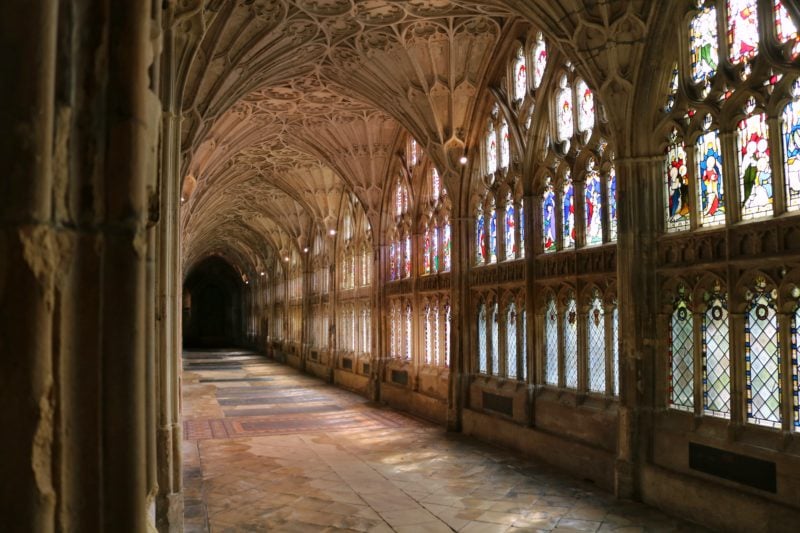 looking down an empty cloister in Gloucester Cathedral with a carved arched cieling built from beige stone and sunlight streaming thorugh stained glass windows. gloucester and cheltenham weekend guide.