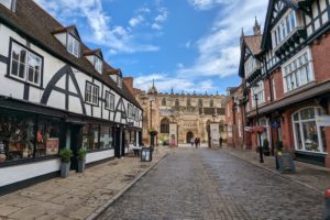 looking down a street with a white tudor style building on the left and red brick buildigns on the right, both containing shops, with part of a cathedral visible at the end of the street