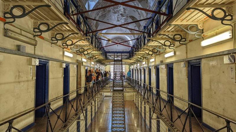 looking down the centre of an abandoned prison with white walls and blue cell doors in rows