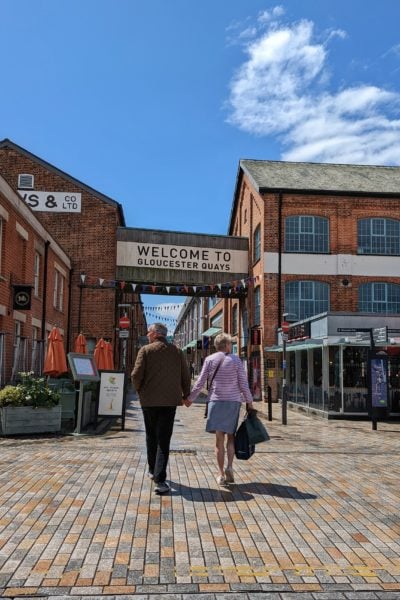 couple walking hand in hand towards a red brick shopping centre labelled Gloucester Quays