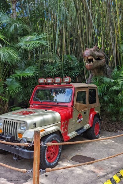model of a predator dinosaur emerging from a thicket of bamboo behind a red and gold Jurassic Park jeep 