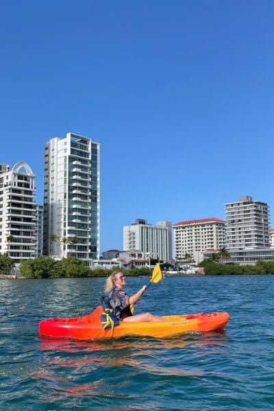 emily wearing a blue shawl with a yellow pattern, sitting on a bright orange kayak in a blue lagoon with tall white tower blocks against a blue sky in san juan puerto rico