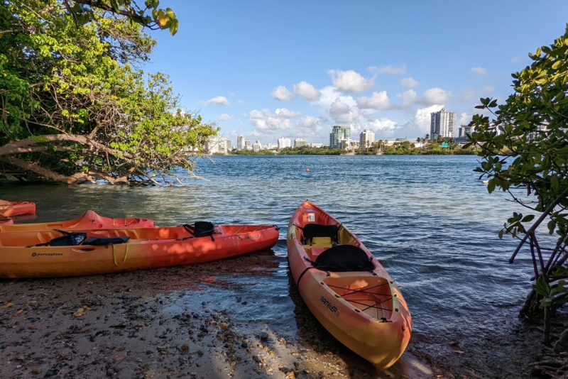 Two orange kayaks on the edge of a large blue lagoon framed by tropical trees with city skyscrapers and tower blocks on the far side of the water - fun things to do in san juan puerto rico