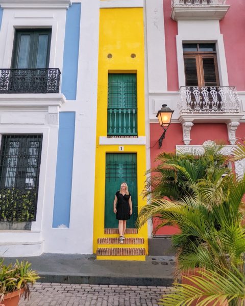 emily wearing a short black dress and converse walking down some steps in front of a dark green door in a very narrow yellow house in san juan puerto rico - simple summer city break packing list! 