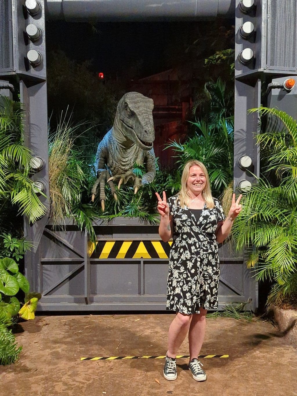 emily wearing a black and white dress standing in front of a metal enclosure with an animatronic velociraptor behind her -  how to do universal studios orlando on a budget