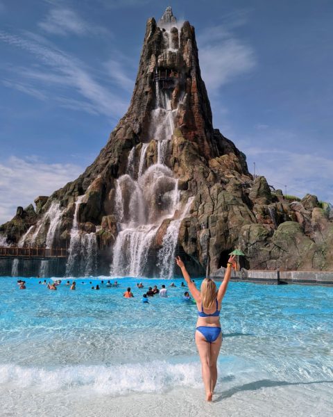 Emily wearing a bright blue bikini facing away from the camera in front of a large blue pool with a giant model volcano behind - Volcano Bay and Universal Studios Orlando on a budget