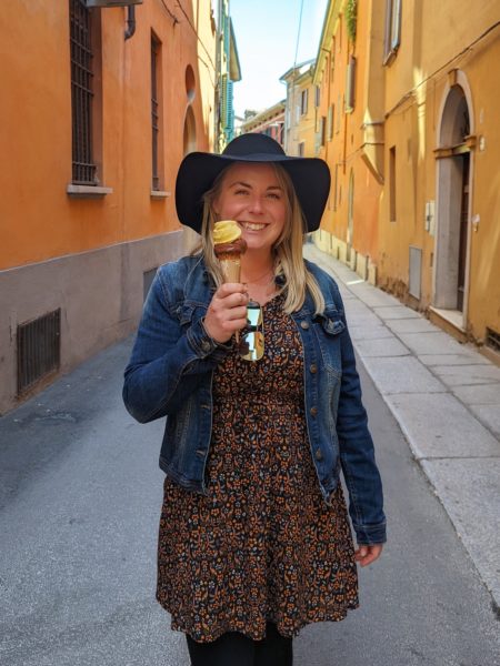 emily in an orange and black dress and denim jacket and black hat holding a yellow ice cream on a cone standing in an alleyway in bologna with orange and yellow buildings on either side.
