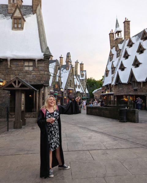 Emily wearing a black and white dress and a Hogwarts robe holding a butterbeer in one hand and a wand in the other standing on a street in Hogsmeade Village at Islands of Adventure, with stone cottages on either side topped with snow -  how to do universal studios orlando on a budget