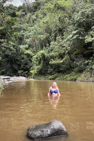 emily wearing a blue bikini top standing in a brown river with the water up to above her waist and many rainforest trees in the background