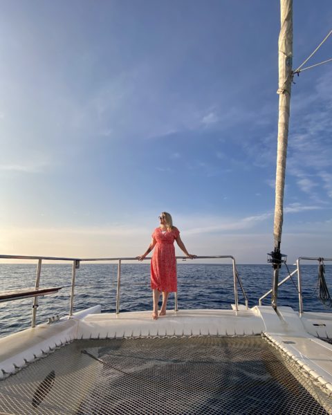 Emily di katamaran di Sri Lanka dengan laut dan langit biru di belakangnya.  Dia mengenakan gaun midi merah cerah dengan bagian depan. 