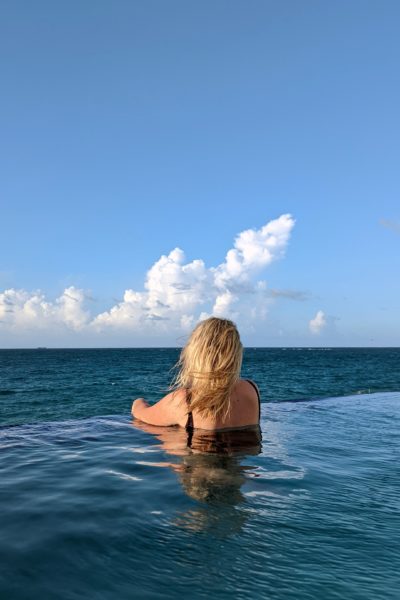 Emily with her back to the camera in an infinity pool facing the ocean on a sunny day with a blue sky above