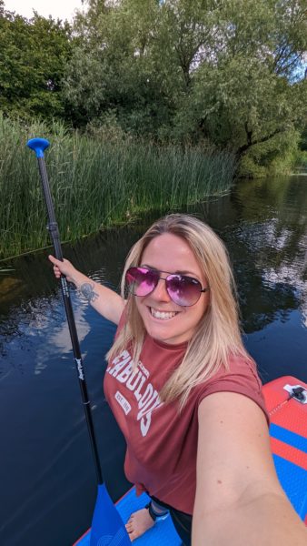 Emily wearing a dark pink top which says FABULOUS in white and pink sunglasses, taking a selfie whilst standing on a paddleboard on a river and holding an oar in her other hand
