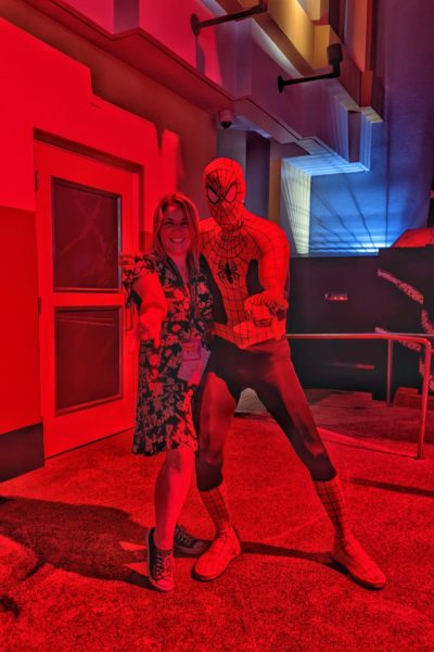 emily wearing a black and white dress at night standing next to Spiderman and pretending to shoot webs from her hand. they are both lit by a red light .