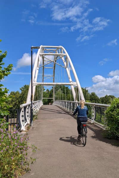 Emily wearing a denim jacket cycling a bike along a paved track towards a white bridge under a blue sky - cheltenham and gloucester weekend