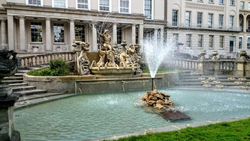 a fountain in front of a gold statue of nelson in front of a white collonaded building in cheltenham city centre