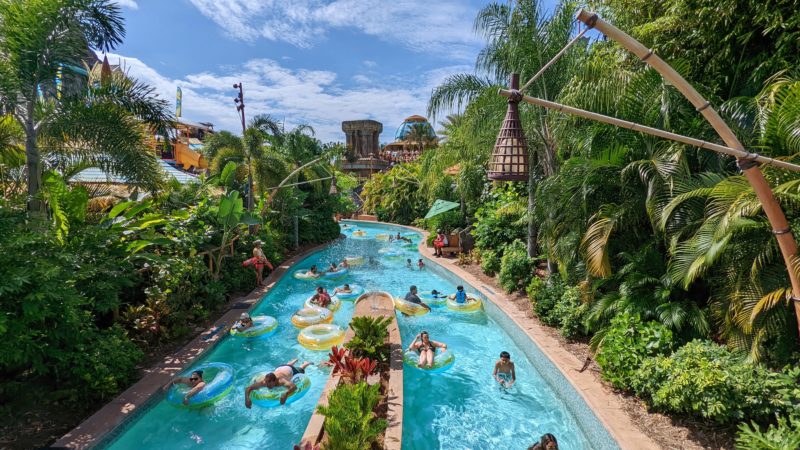 many people on rubber rings in a lazy river with bright blue water between lots of tropical green plants on a very sunny day at Volcano Bay Universal Florida