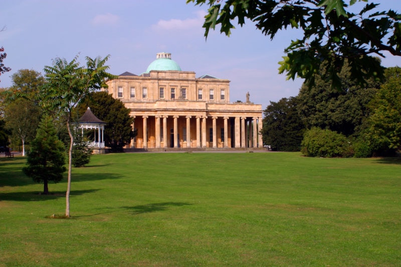 a large green lawn surroudned by trees with a beige coloured classic style building in the centre with many columns along the ground floor - weekend in gloucester and cheltenham