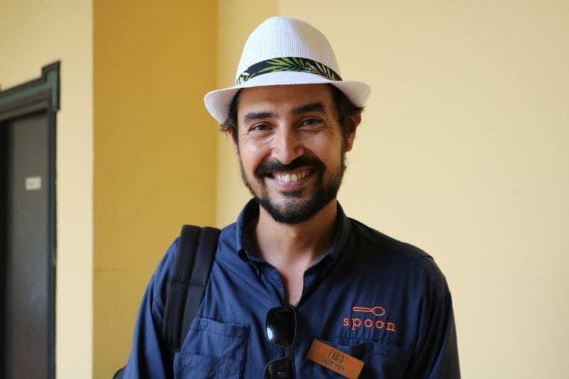 a man with dark hair and a small beard wearing a blue shirt and a straw trilby hat smiling at the camera