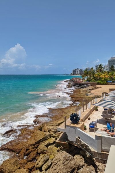 looking down at a terrace with umbrellas and tables next to a rocky beach and turquoise sea with palm trees in the background at Condado Vanderbilt Hotel San Juan Puerto Rico