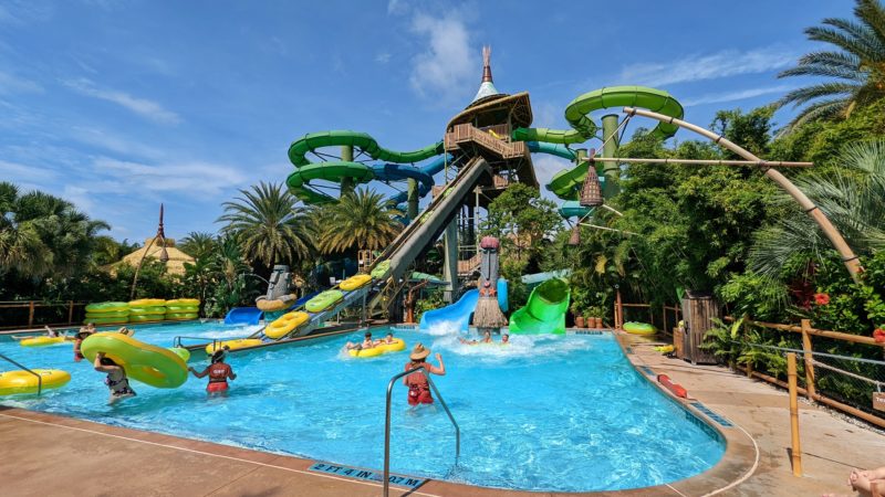 green water slides curling around above a small pool of bright blue water surrounded by palm trees and tropical greenery on a sunny day at Volcano Bay Universal Florida