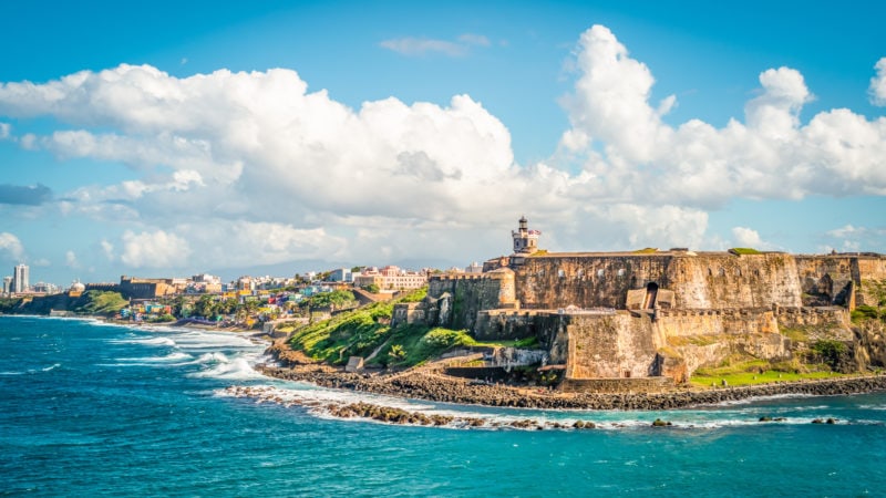 Panoramic landscape of historical castle El Morro - a beige coloured stone fortress with a tower - along the coast in Old San Juan with a city in the background and a bright blue sea in front. fun and unique things to do in puerto rico. 